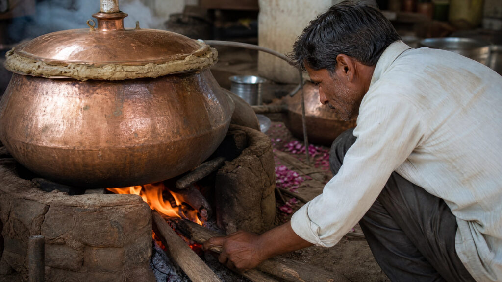 An Indian distiller tends a wood fire beneath a sealed copper deg filled with botanical material during traditional attar production